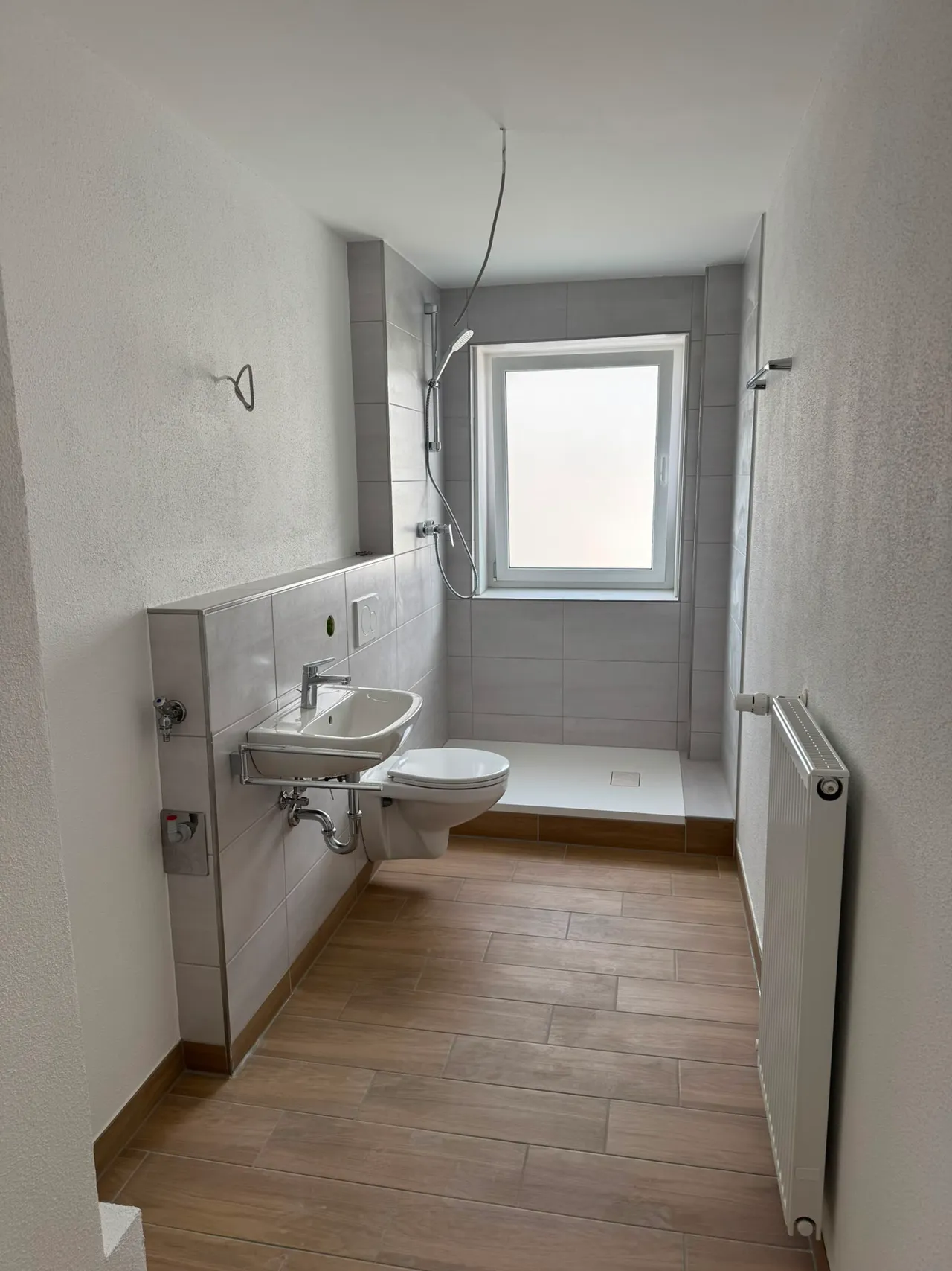Fertiges Bad mit wandhängendem Waschbecken, WC und bodengleicher Dusche unter dem Fenster