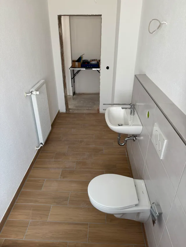 Blick auf WC und Waschbecken vor grau gefliester Vorwand, gegenüber der neue Heizkörper