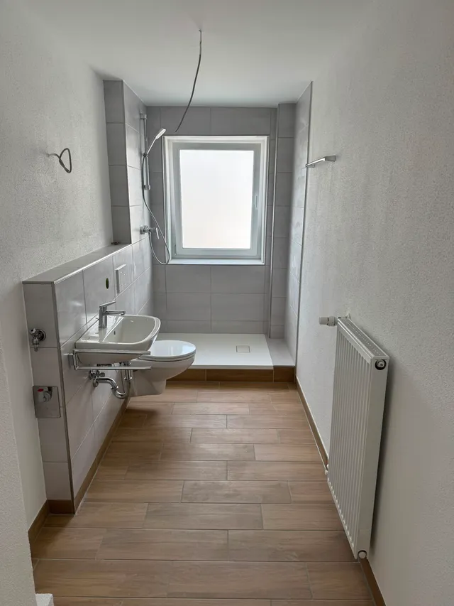 Bodengleiche Dusche mit flacher Duschwanne und Handbrause, daneben der fertig angeschlossene Heizkörper